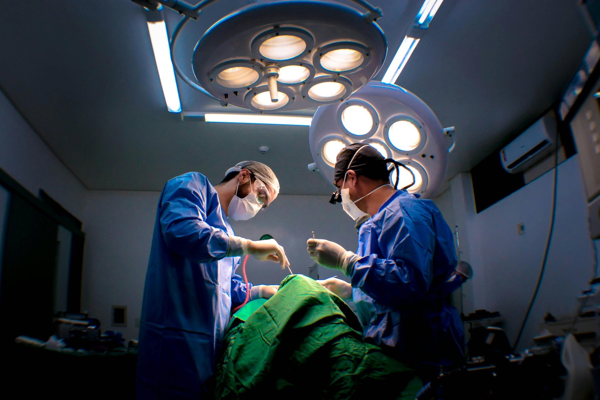 Two surgeons in blue scrubs performing surgery under bright overhead surgical lights in an operating room